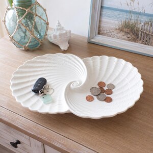 May include: A white ceramic double-bowl dish shaped like two conjoined scallop shells. The dish features a ribbed, radiating texture and a central spiral. It contains a black car key fob with a silver keyring and several copper-colored coins.