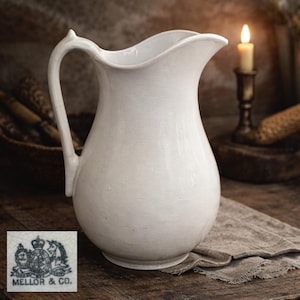 May include: A white ceramic pitcher with a curved handle and a spout. The pitcher has a textured surface and a wide base. A small, blue and white emblem is visible at the bottom left. A lit candle is in the background.