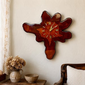 May include: A wall clock crafted from a polished, irregular slab of dark reddish-brown wood. The clock face features gold-colored Roman numerals and matching gold hands. The natural, organic shape of the wood grain is visible throughout the piece.