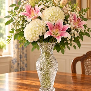 May include: A clear, cut-glass vase with a flared rim and a textured, geometric diamond pattern, holding a large floral arrangement of pink lilies, white hydrangeas, and small white baby's breath flowers with green foliage.