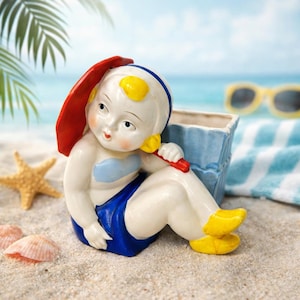 May include: A ceramic figurine of a child in a swimsuit, holding a red parasol. The figurine is white with blue and yellow accents, sitting on a sandy beach with a blue ocean backdrop. A starfish and seashells are nearby.