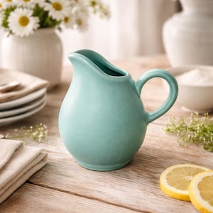 May include: A turquoise ceramic creamer with a curved handle and spout. The creamer is on a wooden surface, with a stack of plates, a vase of flowers, and lemon slices in the background.