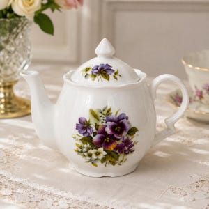 May include: A white porcelain teapot featuring a scalloped rim and a matching lid with a pointed finial. The body and lid are decorated with a vibrant floral pattern of purple and yellow pansies with green leaves, finished with a curved handle.