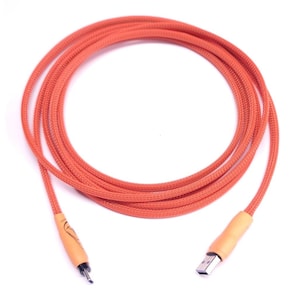 May include: Orange braided USB cable with a light brown connector and a USB-A plug. The cable is coiled and lies on a white surface. The cable has a decorative design on the connector.