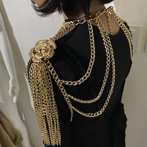 Może przedstawiać: Złota biżuteria na ramię z dużym akcentem róży i kaskadowymi frędzlami z łańcuszków. Element na ramię składa się z wielu łańcuszków i jest noszony na czarnym ubraniu. Biżuteria dodaje odważny, dekoracyjny element.