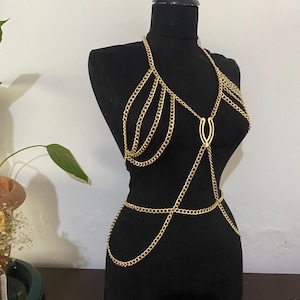 Może przedstawiać: Złoty łańcuszek na ciało z wieloma pasmami i delikatnym wzorem. Łańcuszek jest prezentowany na czarnym manekinie, prezentując jego skomplikowane detale i efekt drapowania. Biżuteria jest przeznaczona do noszenia na klatce piersiowej i wokół talii.