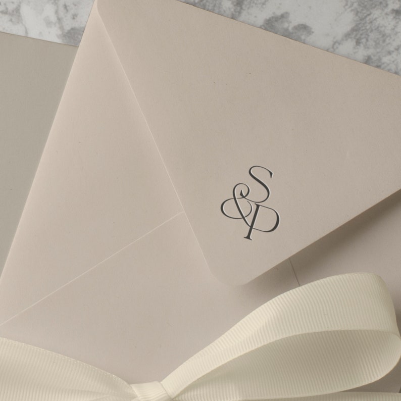 May include: A light beige envelope with a white ribbon tied in a bow. The envelope has a monogram of the letters "S" and "P" in a script font.