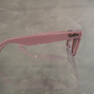 Puede incluir: Gafas de sol Ray-Ban rosas. Las monturas son de un rosa liso y brillante. El logotipo de Ray-Ban es visible en la patilla. Las lentes son de color marr&oacute;n claro. Las gafas de sol est&aacute;n sobre una superficie reflectante.
