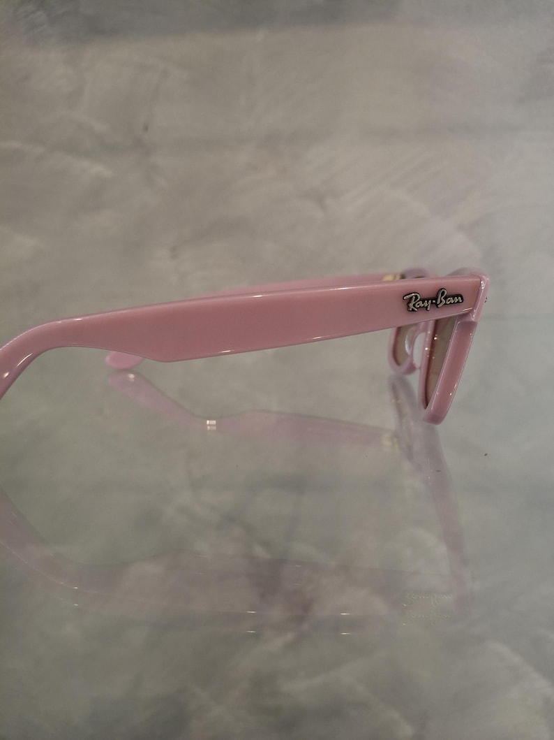 Puede incluir: Gafas de sol Ray-Ban rosas. Las monturas son de un rosa liso y brillante. El logotipo de Ray-Ban es visible en la patilla. Las lentes son de color marr&oacute;n claro. Las gafas de sol est&aacute;n sobre una superficie reflectante.