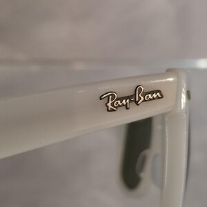 Pu&ograve; includere: Primo piano di occhiali da sole Ray-Ban bianchi. Il logo Ray-Ban &egrave; visibile sull'asta in lettere nere. Gli occhiali da sole hanno una finitura lucida.