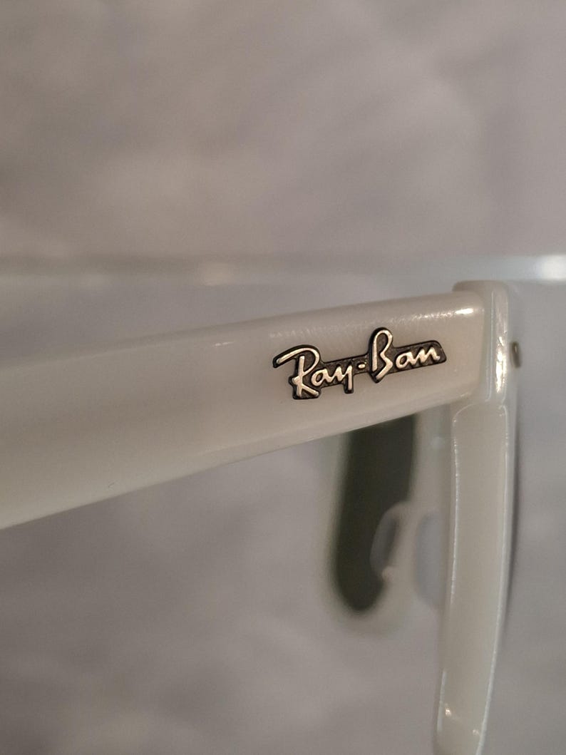Pu&ograve; includere: Primo piano di occhiali da sole Ray-Ban bianchi. Il logo Ray-Ban &egrave; visibile sull'asta in lettere nere. Gli occhiali da sole hanno una finitura lucida.