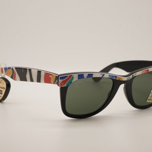 K&ouml;nnte beinhalten: Schwarze Sonnenbrille im Wayfarer-Stil mit gr&uuml;nen Gl&auml;sern und einem bunten, abstrakten Muster auf dem Rahmen. Die Sonnenbrille hat ein Ray-Ban-Logo auf der rechten Linse.