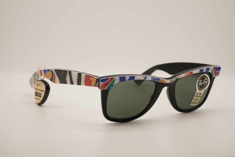 K&ouml;nnte beinhalten: Schwarze Sonnenbrille im Wayfarer-Stil mit gr&uuml;nen Gl&auml;sern und einem bunten, abstrakten Muster auf dem Rahmen. Die Sonnenbrille hat ein Ray-Ban-Logo auf der rechten Linse.