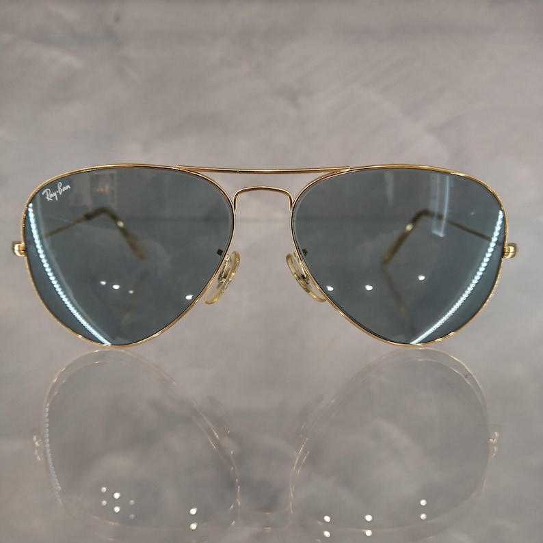 May include: Gold-framed aviator sunglasses with blue-tinted lenses. The Ray-Ban logo is visible on the upper left lens. The sunglasses are reflected on a shiny surface.