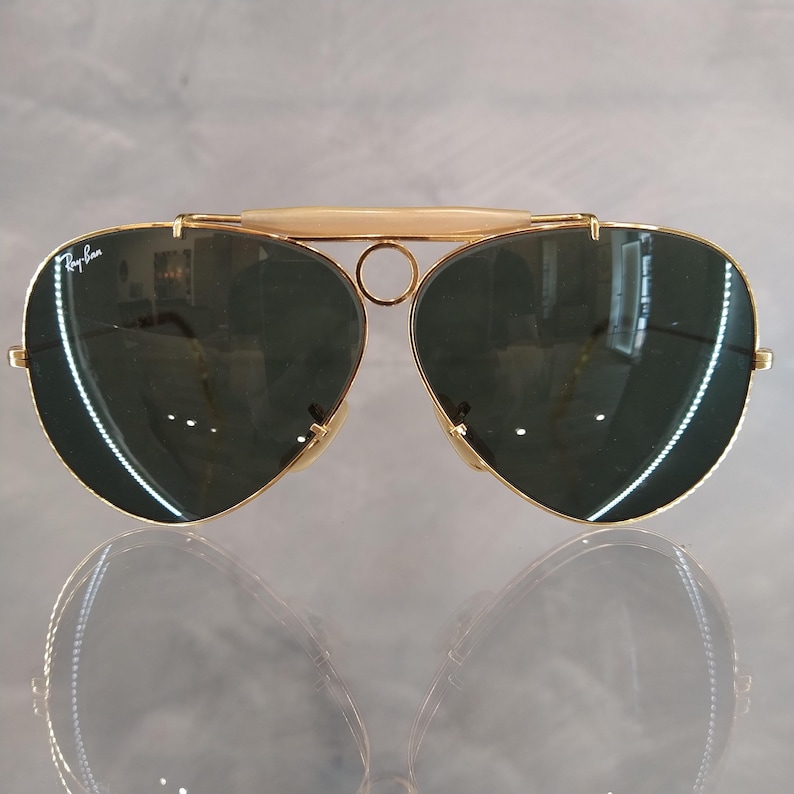 Puede incluir: Gafas de sol aviador doradas con lentes verde oscuro y montura de metal dorado. Las gafas tienen un dise&ntilde;o distintivo de doble puente.