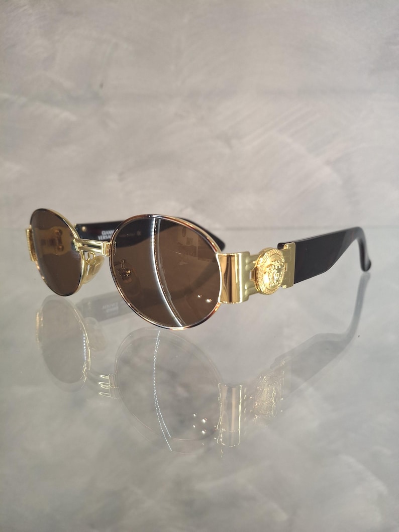 K&ouml;nnte beinhalten: Sonnenbrille mit goldfarbenem Rahmen und braunen Gl&auml;sern. Die Sonnenbrille hat ein goldenes Emblem an den B&uuml;geln und schwarze B&uuml;gel. Die Gl&auml;ser sind rund und spiegeln die Umgebung wider. Die Sonnenbrille liegt auf einer reflektierenden Oberfl&auml;che.