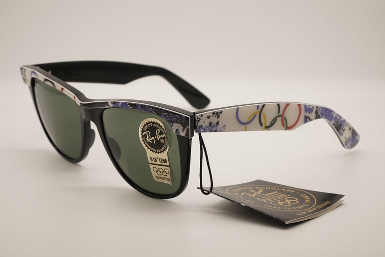 Puede incluir: Gafas de sol Ray-Ban negras con lentes verdes y un patr&oacute;n de anillos ol&iacute;mpicos blancos, azules y rojos en las patillas. Las gafas de sol vienen con una etiqueta negra que dice "Ray-Ban Sunglasses 1984 Olympic Games"