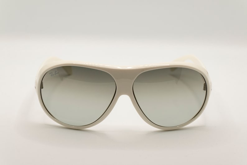 Puede incluir: Gafas de sol color crema con lentes degradadas. Las gafas tienen una montura gruesa y un dise&ntilde;o curvo. El logotipo de Ray-Ban es visible en la lente superior izquierda. Las lentes pasan de un tono m&aacute;s oscuro en la parte superior a uno m&aacute;s claro en la inferior.