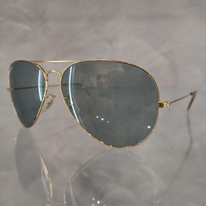 Puede incluir: Gafas de sol aviador doradas con lentes grises oscuros. Las gafas est&aacute;n sobre una superficie blanca.