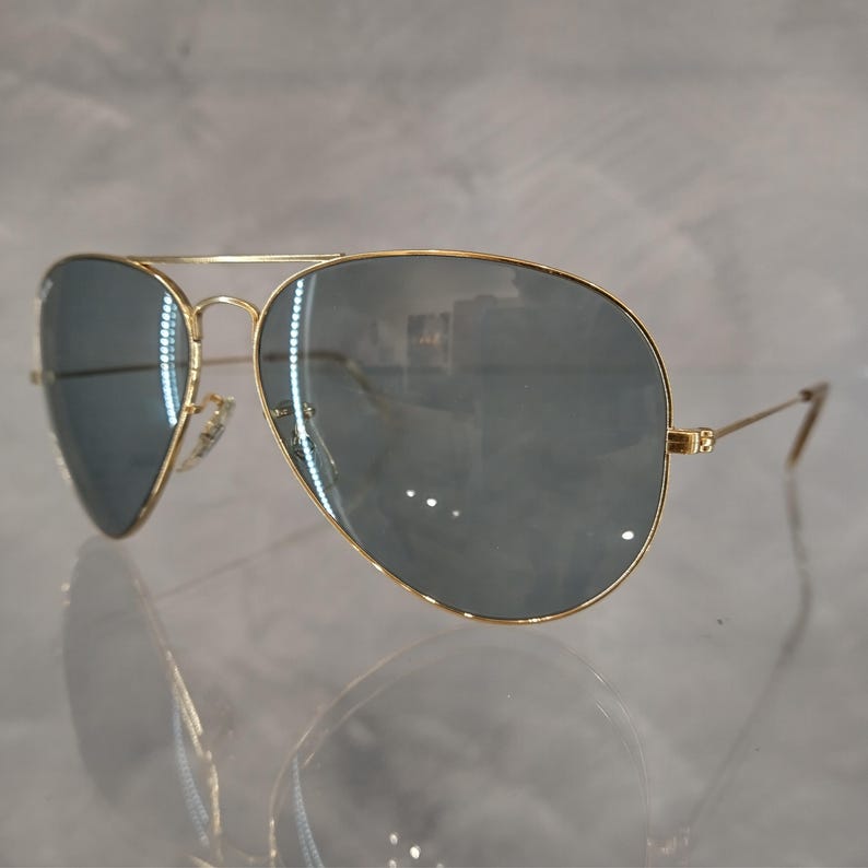 Puede incluir: Gafas de sol aviador doradas con lentes grises oscuros. Las gafas est&aacute;n sobre una superficie blanca.