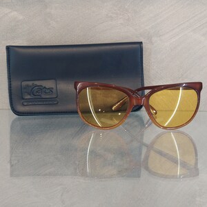 May include: Brown cat-eye sunglasses with yellow lenses and a black leather case with the text "Ray-Ban Cats 14K Goldtone" printed on it.