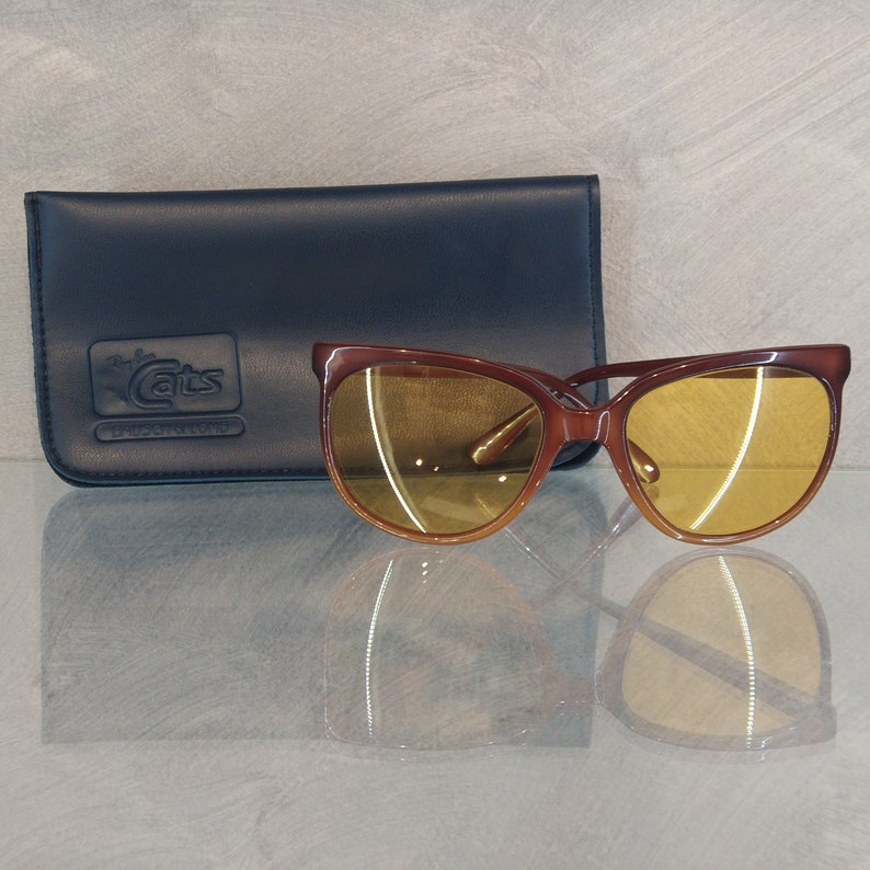 May include: Brown cat-eye sunglasses with yellow lenses and a black leather case with the text "Ray-Ban Cats 14K Goldtone" printed on it.