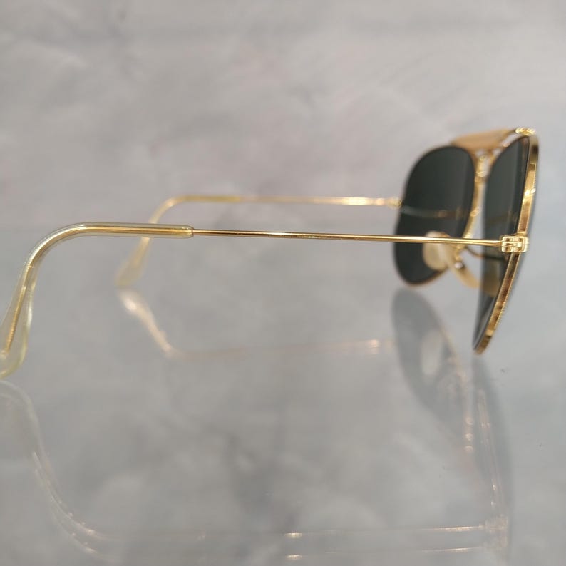 Puede incluir: Gafas de sol aviador doradas con lentes verde oscuro. Las gafas tienen un fino marco de metal y se muestran de lado.