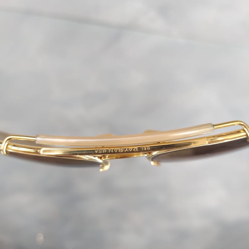 May include: Close-up of the gold-tone metal frame of a pair of sunglasses. The frame has a brown plastic nose bridge and the text "B&L RAY-BAN USA" is engraved on the frame.