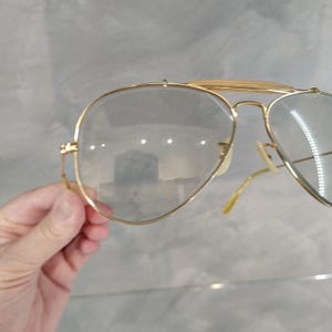 May include: Gold aviator sunglasses with clear lenses. The sunglasses are held in a hand.