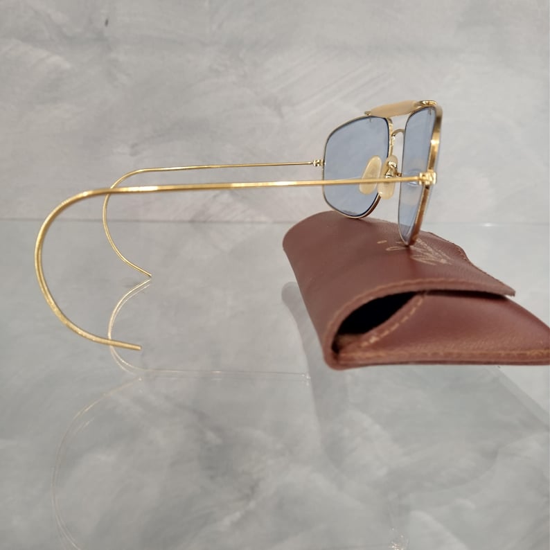Puede incluir: Gafas de sol aviador doradas con lentes azules y una funda de cuero marr&oacute;n. Las gafas tienen un fino marco met&aacute;lico y patillas curvadas de alambre.