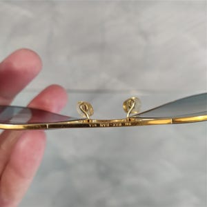 May include: Close-up of gold-rimmed sunglasses with blue-tinted lenses. The frame has a classic design with a thin, rectangular shape. The nose pads are a light gold colour. The text "RAY-BAN USA" is visible on the frame.