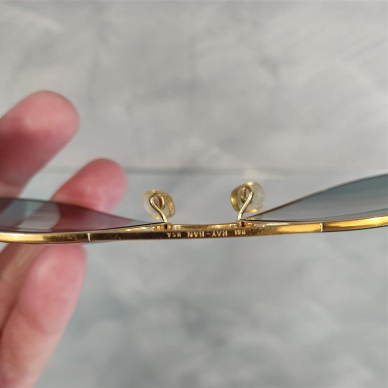 May include: Close-up of gold-rimmed sunglasses with blue-tinted lenses. The frame has a classic design with a thin, rectangular shape. The nose pads are a light gold colour. The text "RAY-BAN USA" is visible on the frame.