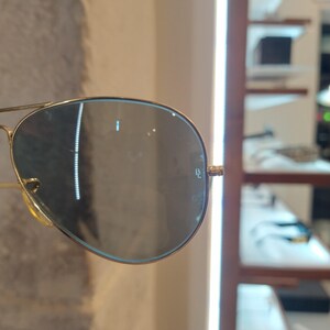 May include: Gold-framed aviator sunglasses with dark lenses. The lenses have a reflective quality, and the frame features a classic double bridge design. The sunglasses are displayed in a store setting.