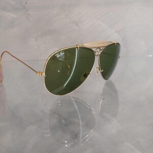May include: Gold-tone aviator sunglasses with green lenses. The sunglasses have a Ray-Ban logo on the left lens.