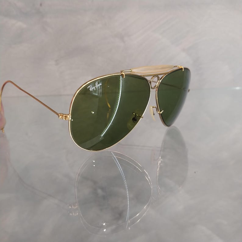 May include: Gold-tone aviator sunglasses with green lenses. The sunglasses have a Ray-Ban logo on the left lens.