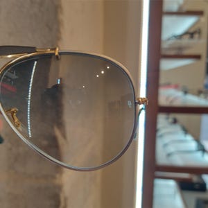 Puede incluir: Primer plano de una gafas de sol aviador con montura dorada y lentes grises. Las gafas reflejan la luz de un expositor de tienda.