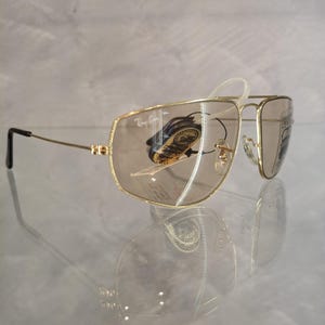 May include: Gold-framed aviator sunglasses with brown tinted lenses. The sunglasses have a double bridge and a classic teardrop shape. The Ray-Ban logo is visible on the top left lens. The sunglasses are displayed on a reflective surface.
