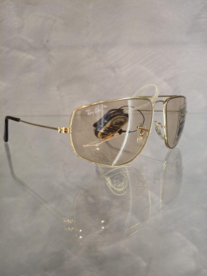 May include: Gold-framed aviator sunglasses with brown tinted lenses. The sunglasses have a double bridge and a classic teardrop shape. The Ray-Ban logo is visible on the top left lens. The sunglasses are displayed on a reflective surface.