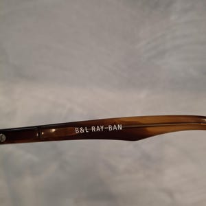 May include: Close-up of a brown and tan Ray-Ban eyeglass arm. The text "B&L RAY-BAN" is printed in white on the arm. The arm has a tortoiseshell pattern.