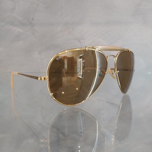 May include: Gold aviator sunglasses with brown lenses. The sunglasses have a metal frame and a bridge with a white plastic insert.