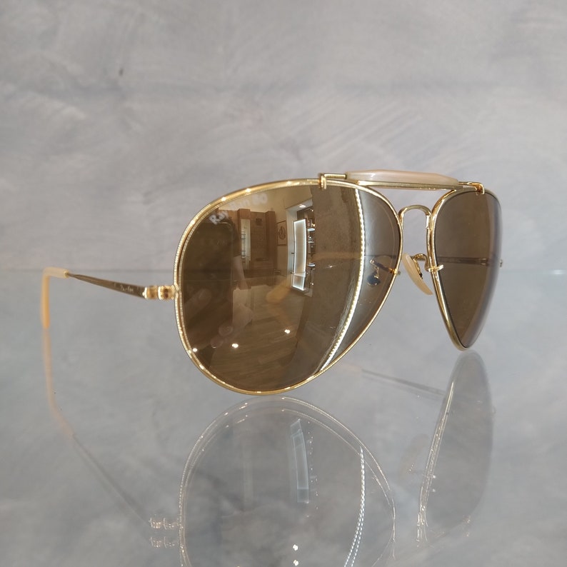 May include: Gold aviator sunglasses with brown lenses. The sunglasses have a metal frame and a bridge with a white plastic insert.