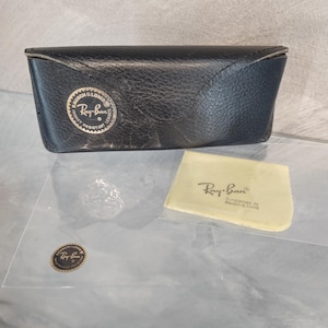 Puede incluir: Un estuche de gafas de sol Ray-Ban de cuero negro con un logotipo dorado en relieve. El estuche est&aacute; cerrado y tiene un peque&ntilde;o pa&ntilde;o de limpieza amarillo con el logotipo de Ray-Ban y el texto "Sunglasses by Bausch & Lomb".