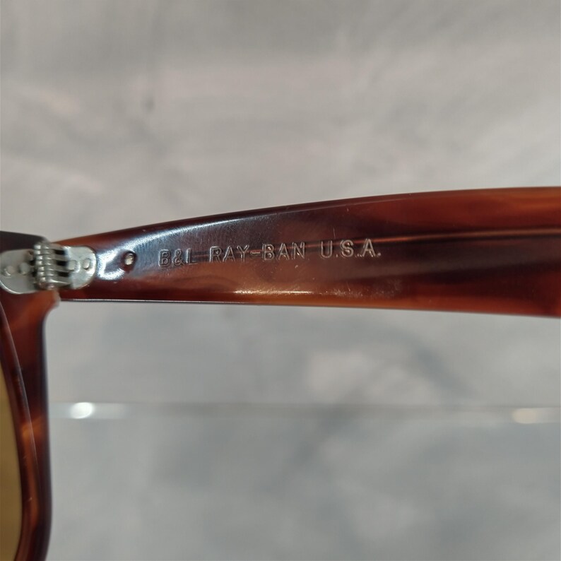 May include: Close-up of the brown tortoiseshell frame of a pair of Ray-Ban sunglasses. The text "B&L RAY-BAN U.S.A." is engraved on the frame.