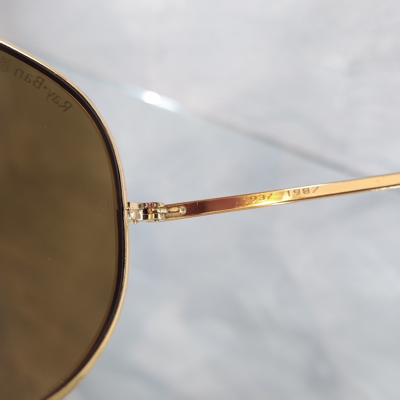 May include: Gold-tone aviator sunglasses with brown lenses. The sunglasses have a Ray-Ban logo on the left arm and the date 1997 1987 on the right arm.