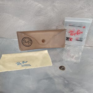Puede incluir: Un estuche de gafas de sol Ray-Ban de cuero marr&oacute;n con cierre de presi&oacute;n y un logotipo circular de Ray-Ban. El estuche viene acompa&ntilde;ado de una peque&ntilde;a bolsa de pl&aacute;stico transparente que contiene un par de gafas de sol Ray-Ban y un pa&ntilde;o de limpieza.
