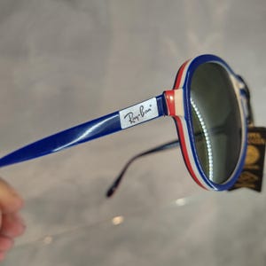 May include: Blue and white Ray-Ban sunglasses with a red stripe on the frame. The sunglasses have green lenses.