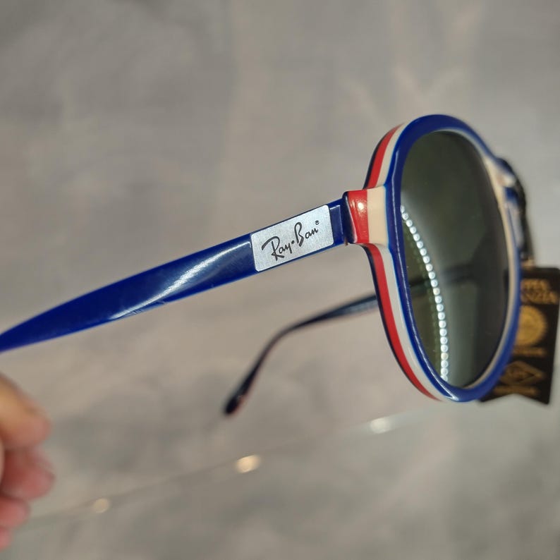 May include: Blue and white Ray-Ban sunglasses with a red stripe on the frame. The sunglasses have green lenses.