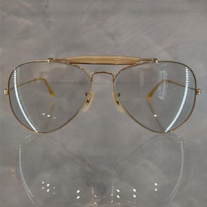 May include: Gold aviator sunglasses with clear lenses. The sunglasses have a thin metal bridge and a gold frame.