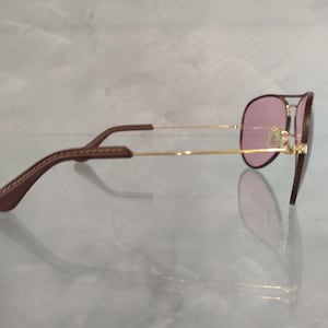 Op de afbeelding: Een gouden aviator zonnebril met bruine leren oorsteunen en roze glazen.
