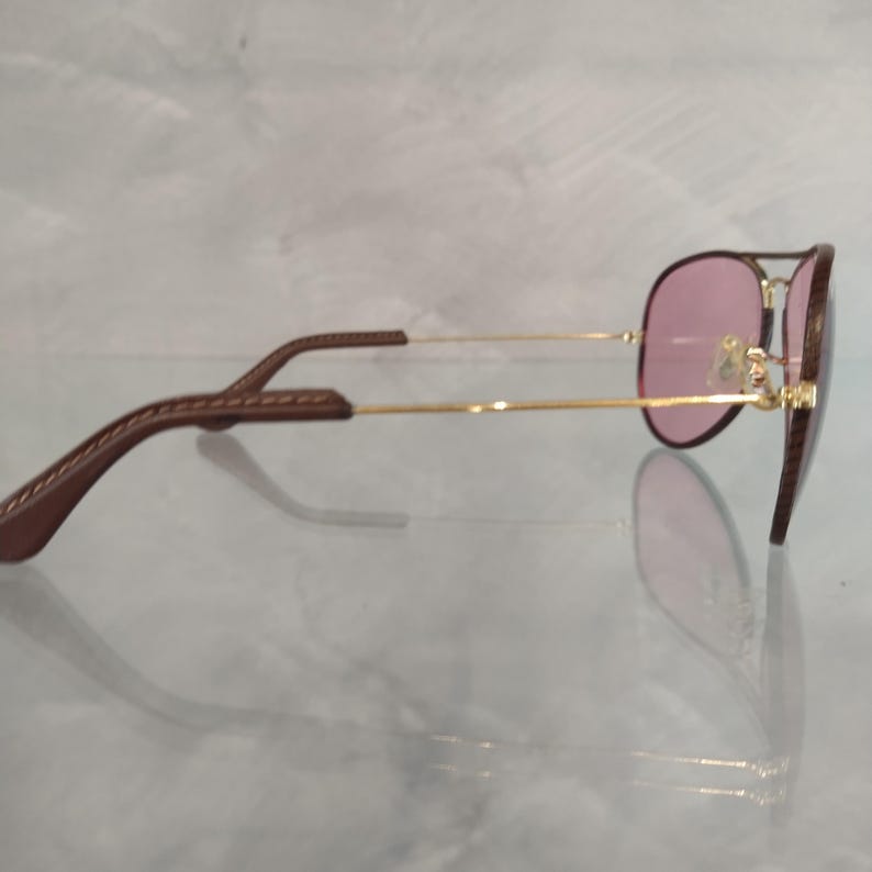 Op de afbeelding: Een gouden aviator zonnebril met bruine leren oorsteunen en roze glazen.
